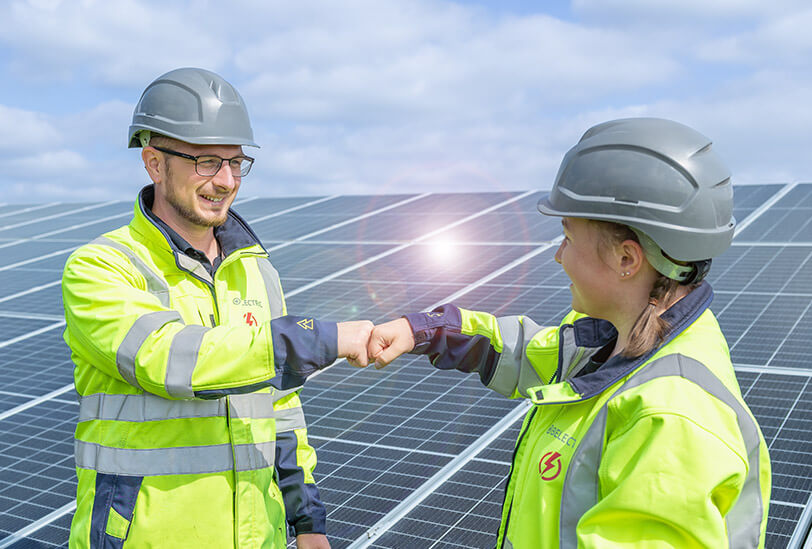 Two electricians who work for a provider of solar systems fist-bump in front of a solar farm