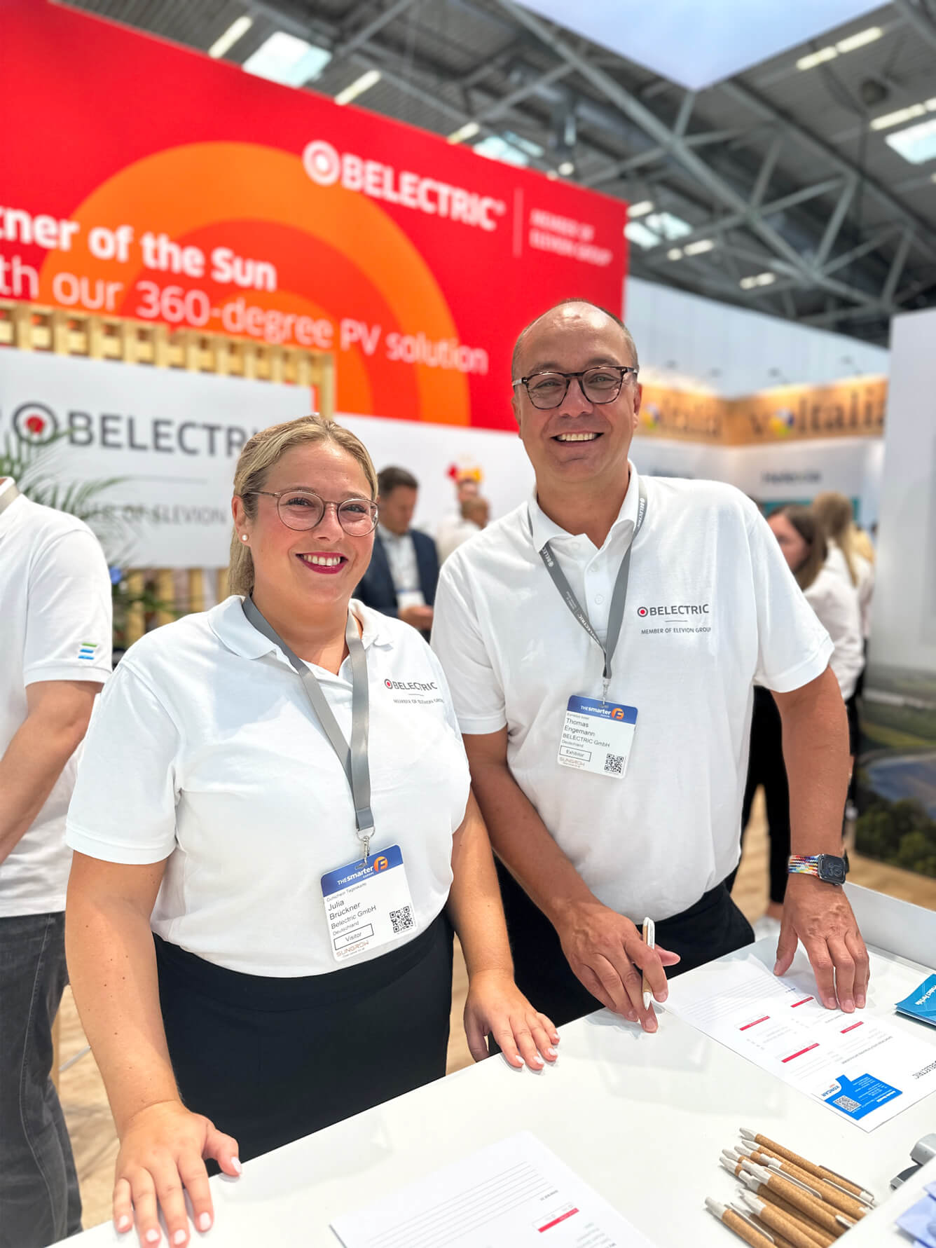 Two employees at one of BELECTRIC's trade fair appearances smile into the camera.