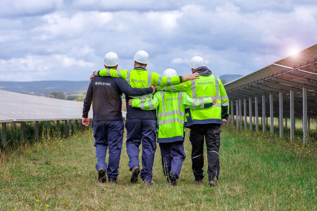 Four BELECTRIC electricians walk across a solar farm, arms around one another