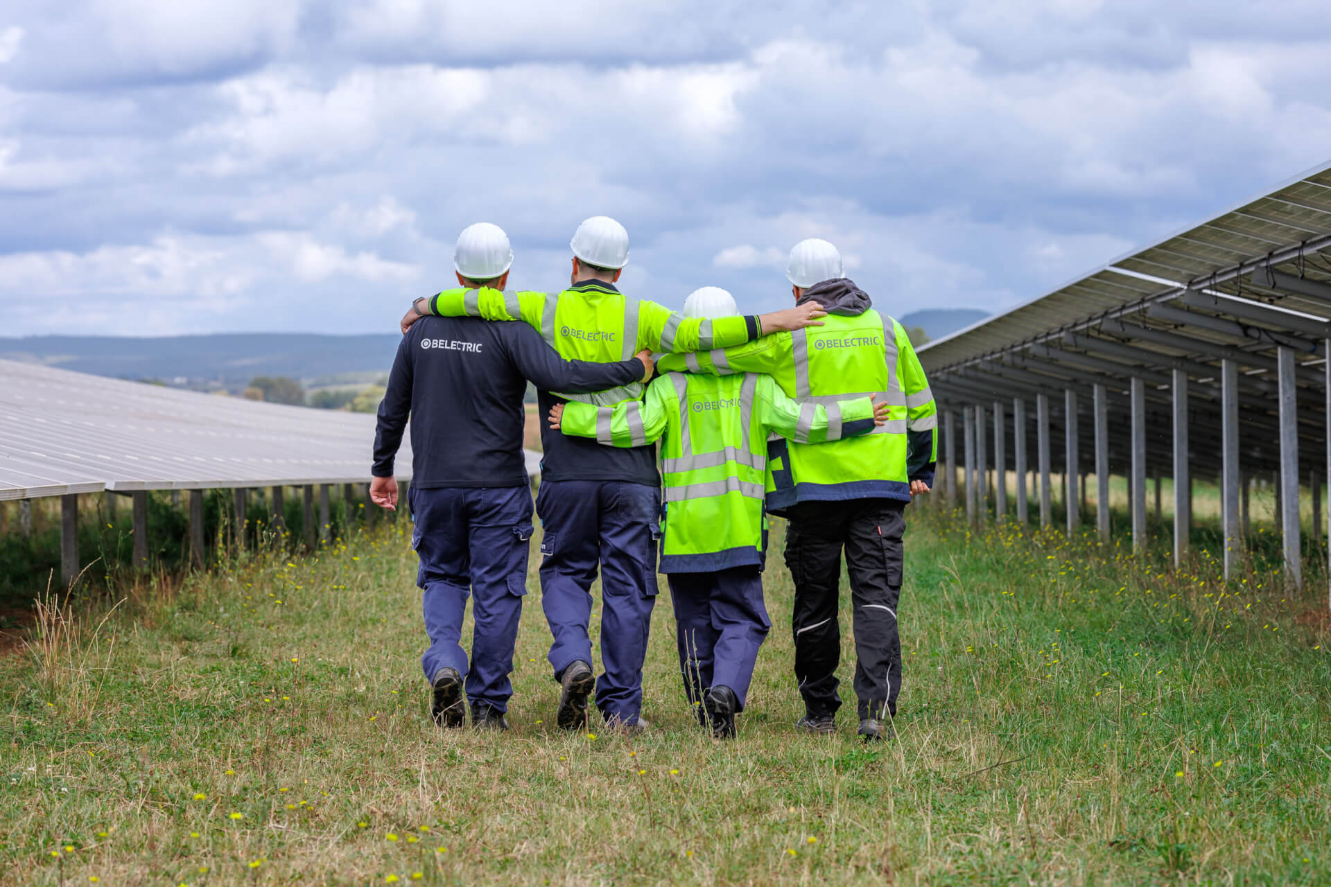 Four BELECTRIC employees in high-visibility jackets walk across a solar farm, arm in arm, celebrating the company's 25th anniversary.